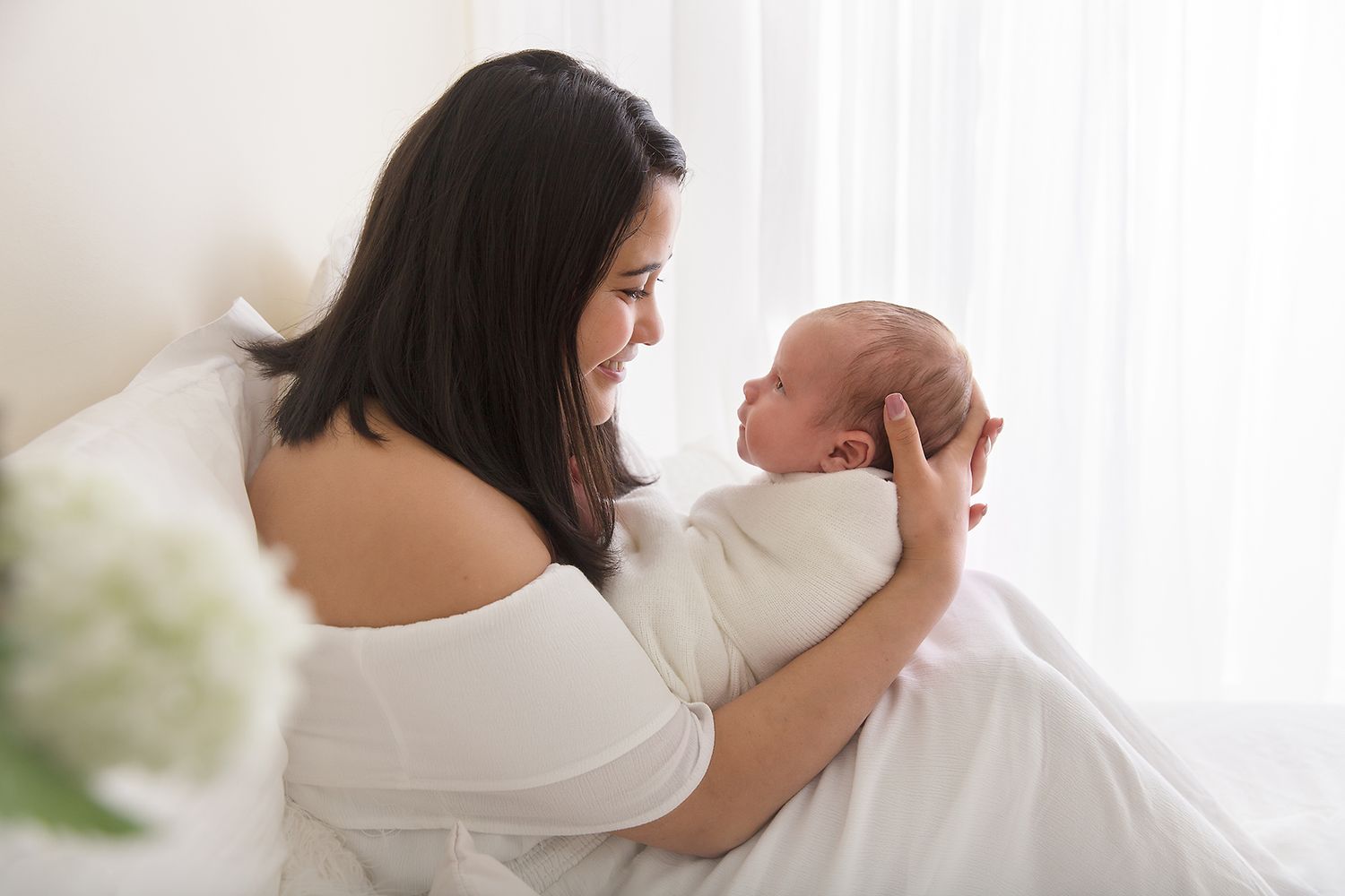 Mother in off-shoulder dress cradling newborn baby near window.