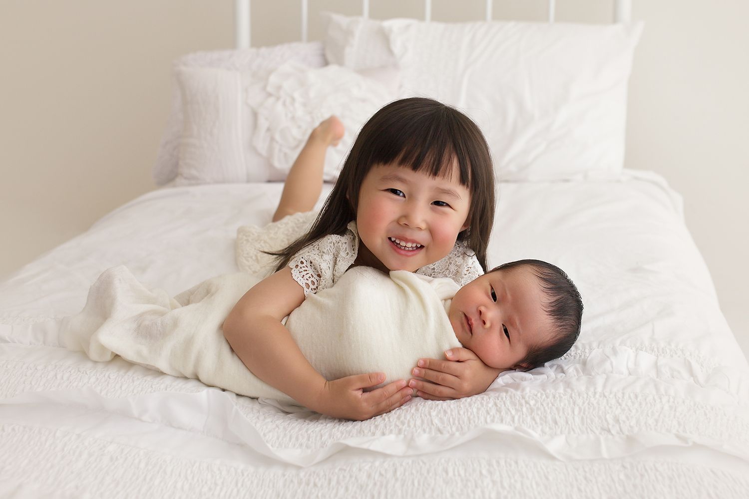 Two siblings on white bedding with older sister embracing newborn baby.