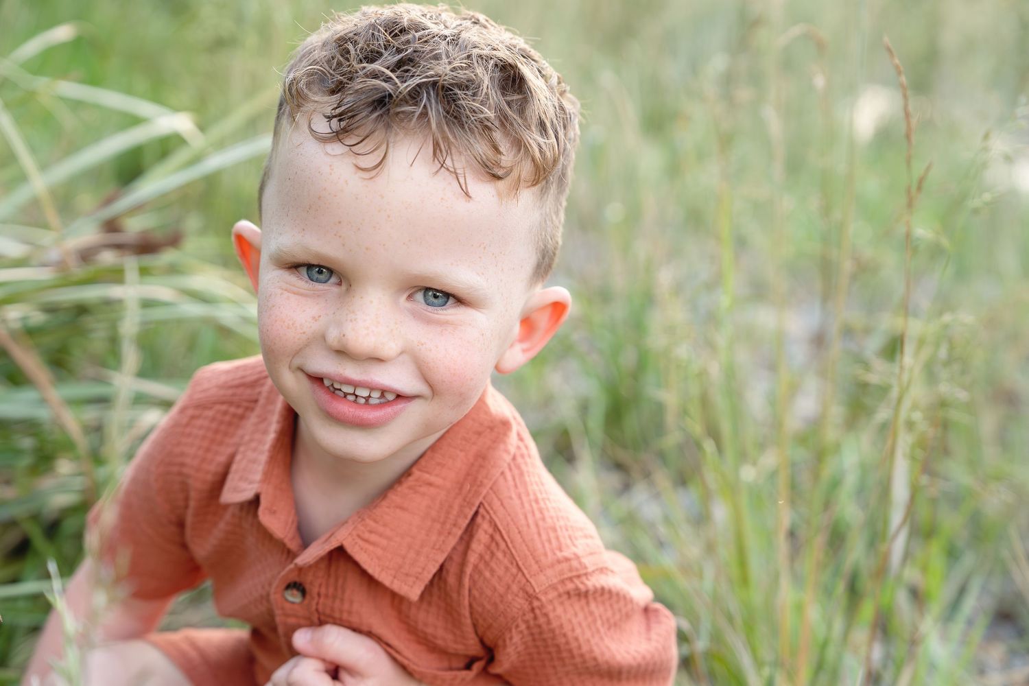 A child in a rust-colored sweater sits in a grassy field on a sunny day.