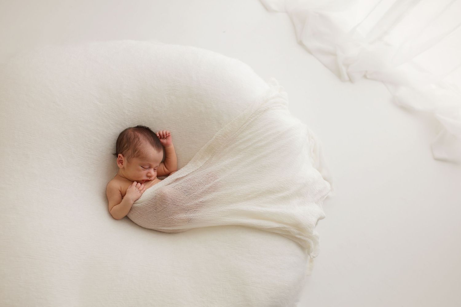 Newborn baby wrapped in soft white fabric against light background.