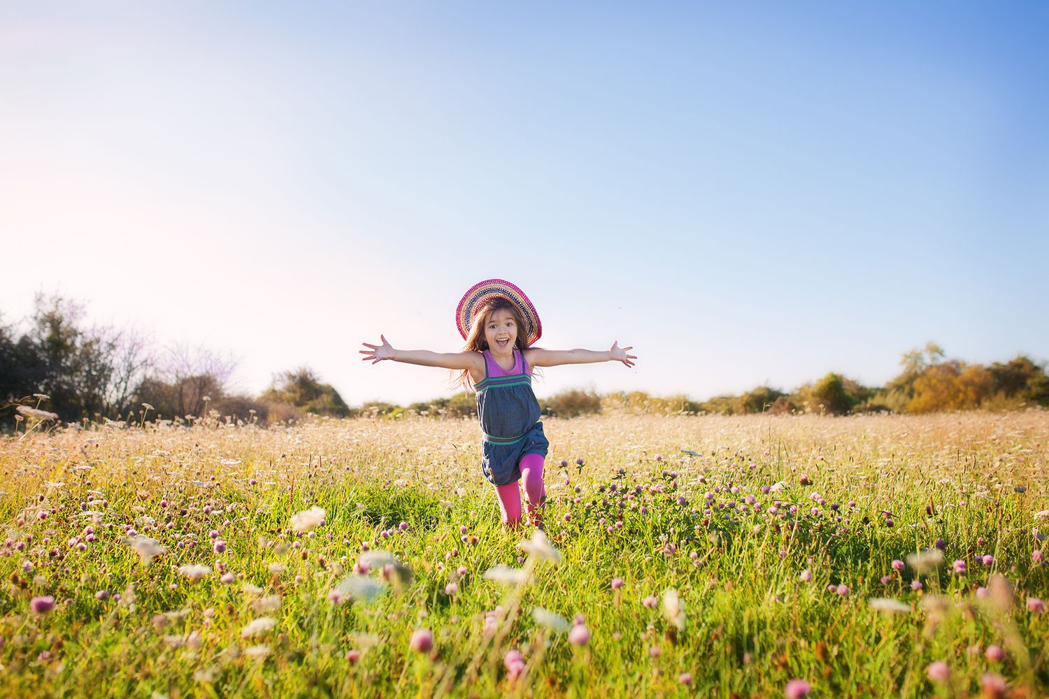 A joyful figure in a blue dress and sunhat jumps with outstretched arms in a wildflower meadow on a sunny day.