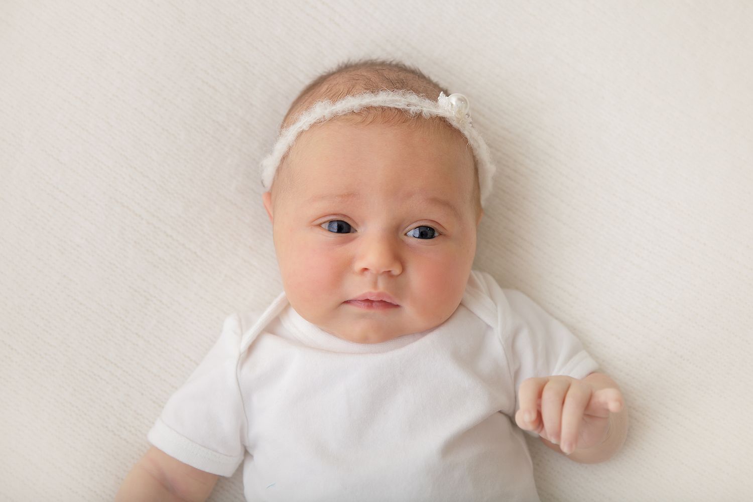 Baby wearing white onesie and headband lying on white background looking at camera.