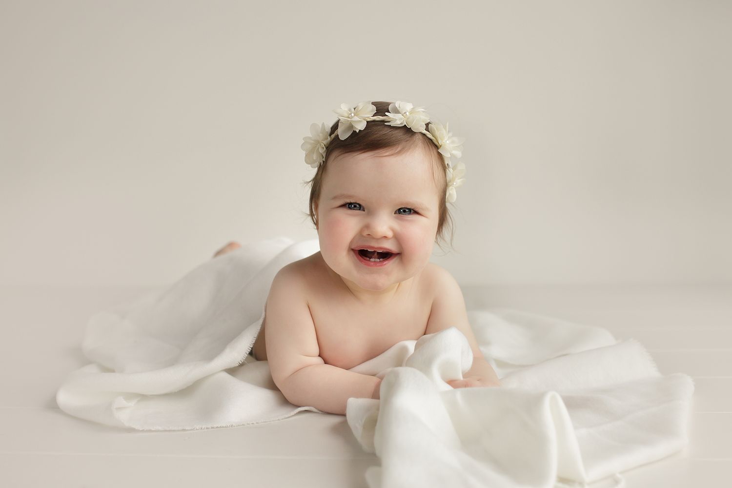 Baby in white outfit smiling while sitting on soft white fabric.