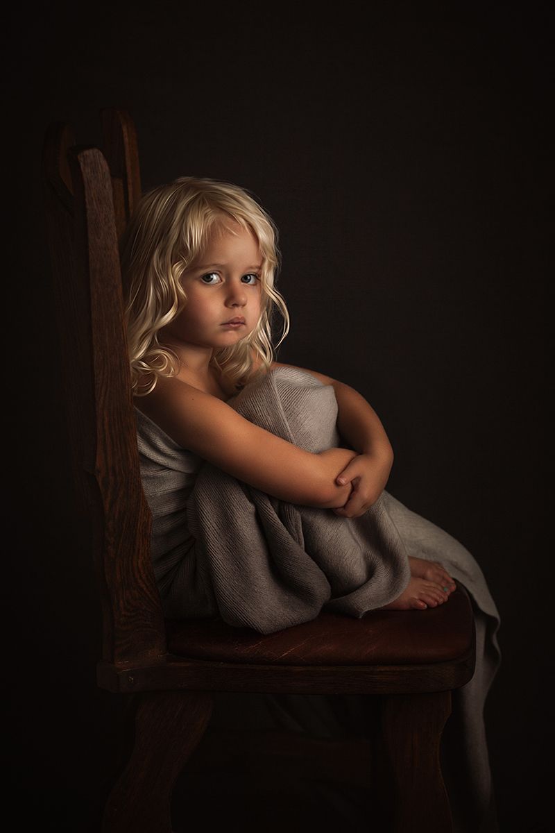 Low key portrait with dramatic shadows showing a figure curled up on a wooden chair.