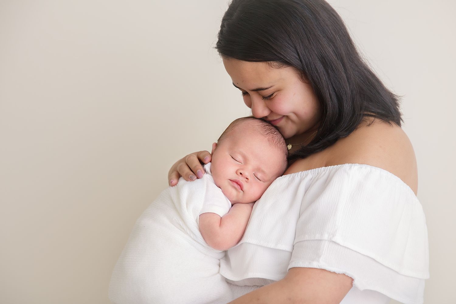 Mother in white off-shoulder dress tenderly embracing sleeping newborn.
