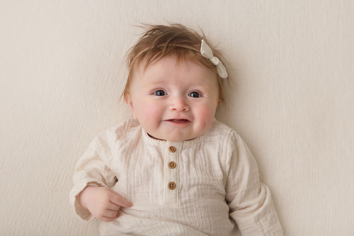 Baby wearing white outfit smiles while lying on white bedding in natural light studio setting.