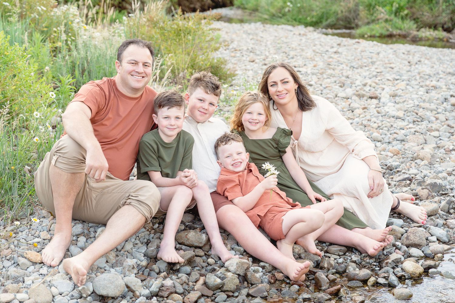 A family of five sits together on a rocky riverbank surrounded by greenery on a sunny day.