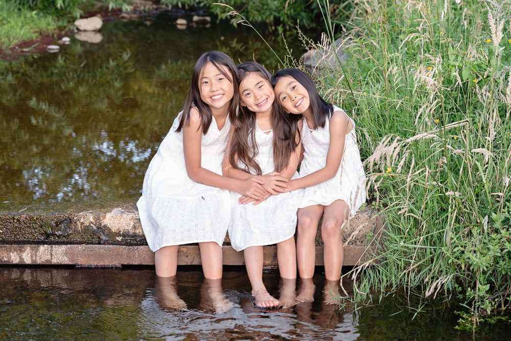 Three people in white dresses sit together at the edge of a pond surrounded by greenery and reeds.