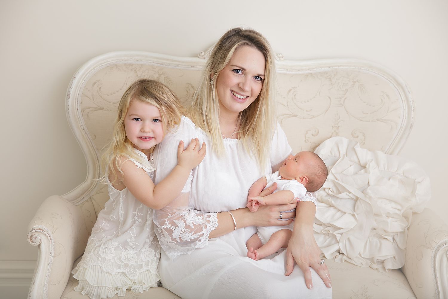 Family portrait on cream chair with newborn and toddler in white dresses.
