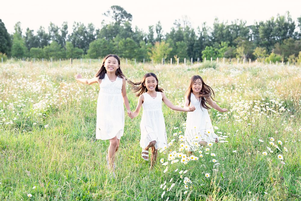 Three people in white dresses walk through a sunlit meadow filled with wildflowers and tall grass.