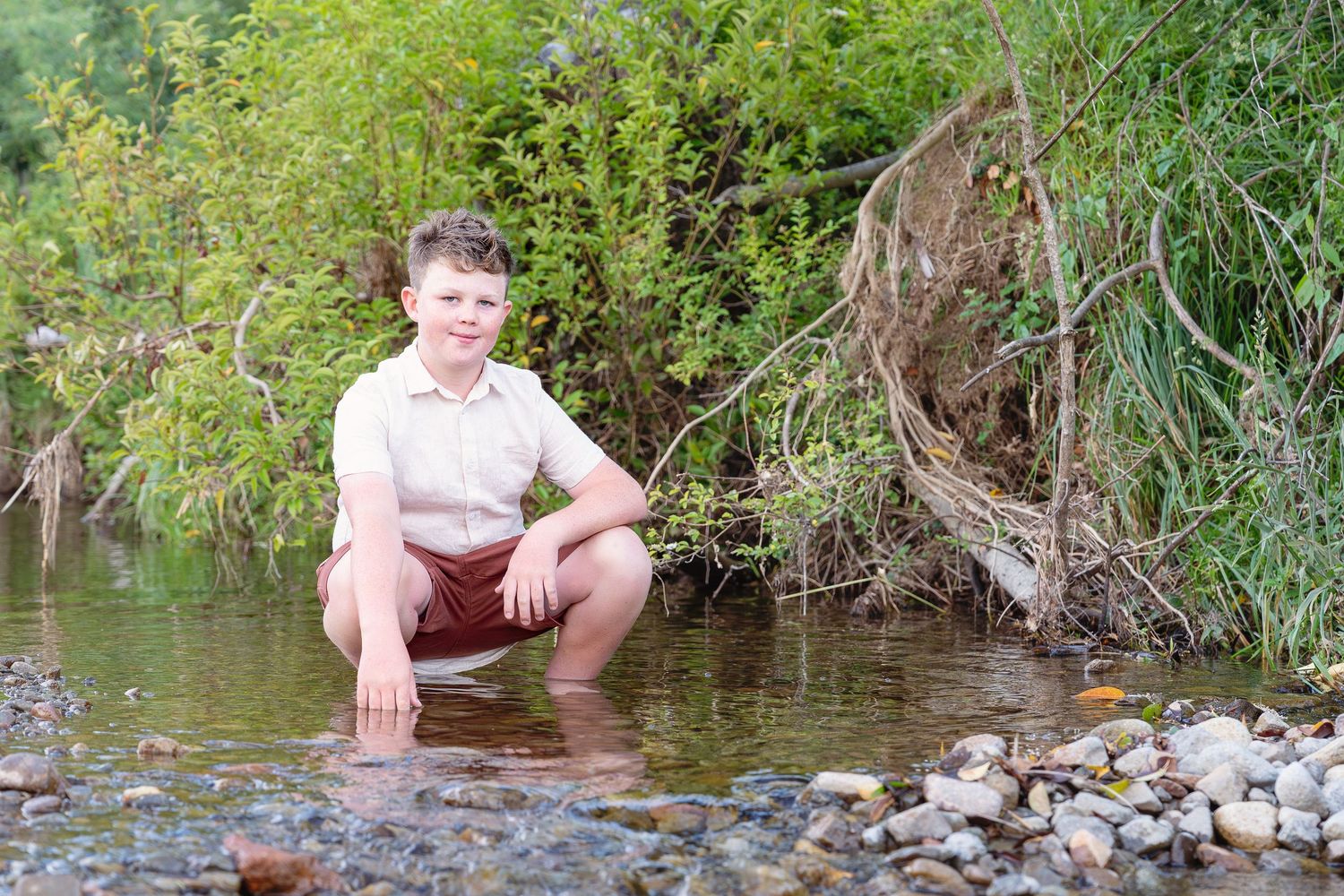 Children splash and play in a shallow creek on a warm summer day while surrounded by greenery and rocks.
