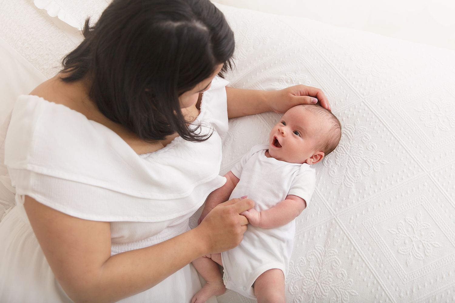 Mother in white dress gently interacting with baby on white bedding.