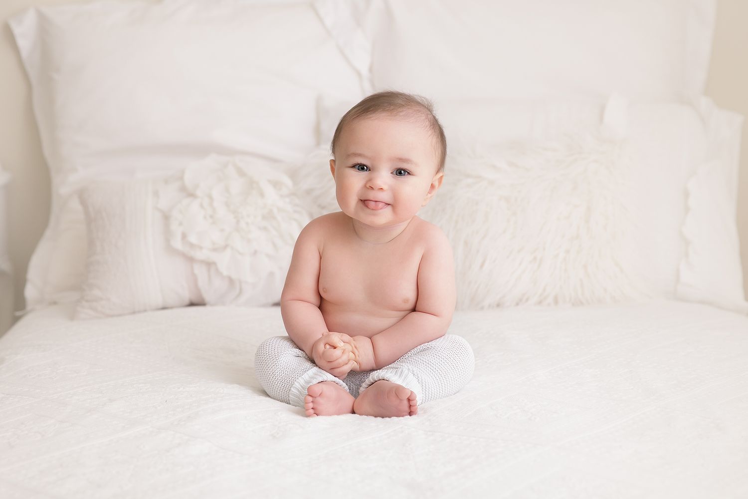 Baby in soft white clothing sits upright on white bedding during professional photo session.
