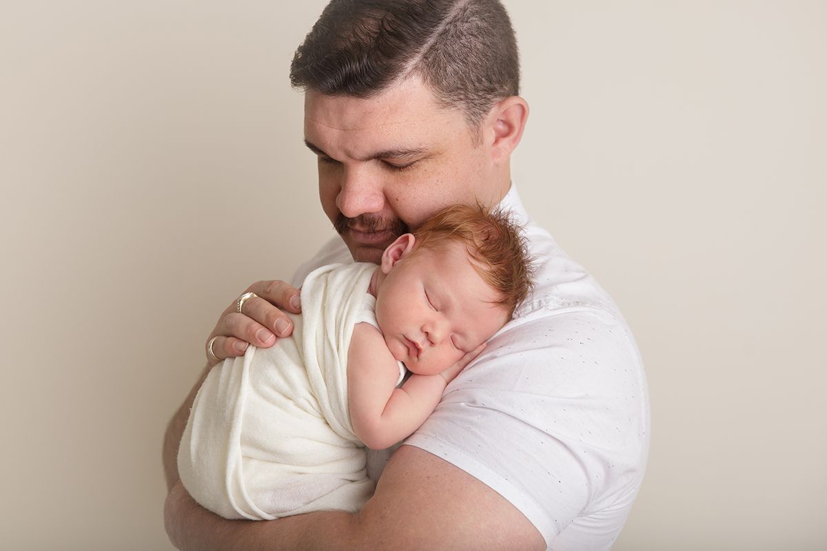 Father in white shirt holding sleeping newborn baby against neutral background.