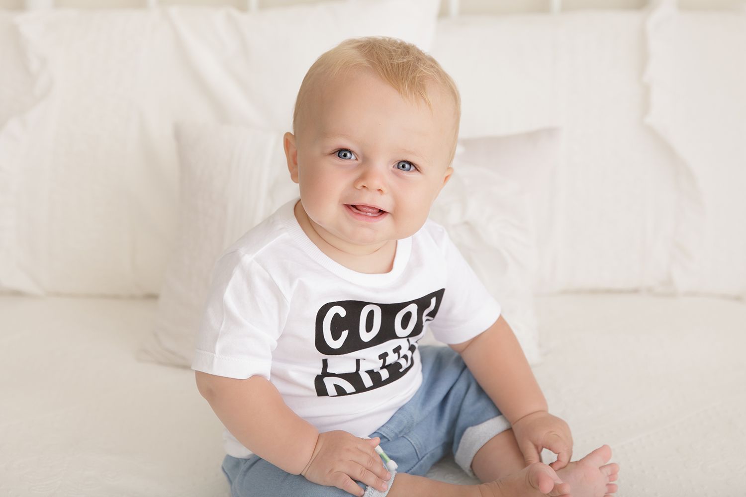 Baby in white t-shirt and blue jeans sits on white background with happy expression.