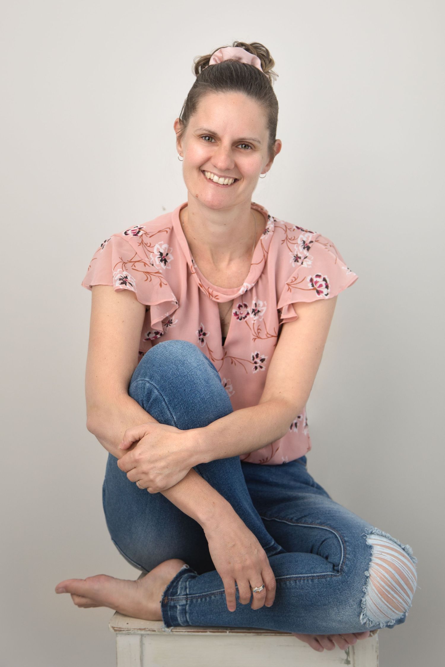 Woman in pink floral top and ripped jeans sitting casually against white studio backdrop.