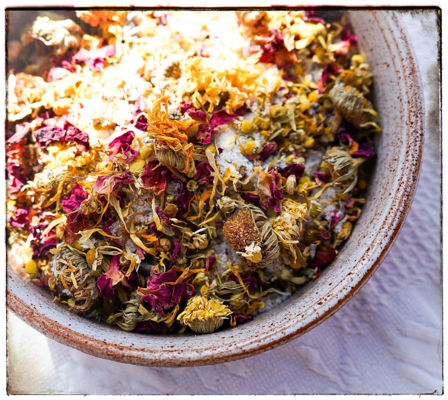 Dried flowers and petals in rustic ceramic bowl with mix of vibrant red and yellow colors on white linens.