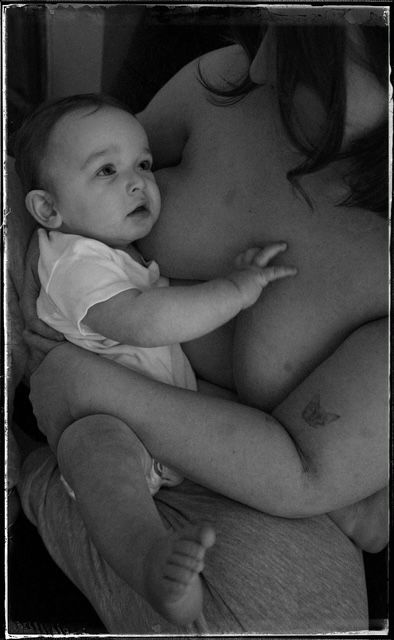 A black and white photograph shows an infant in a white t-shirt reaching up while nestled against someone.