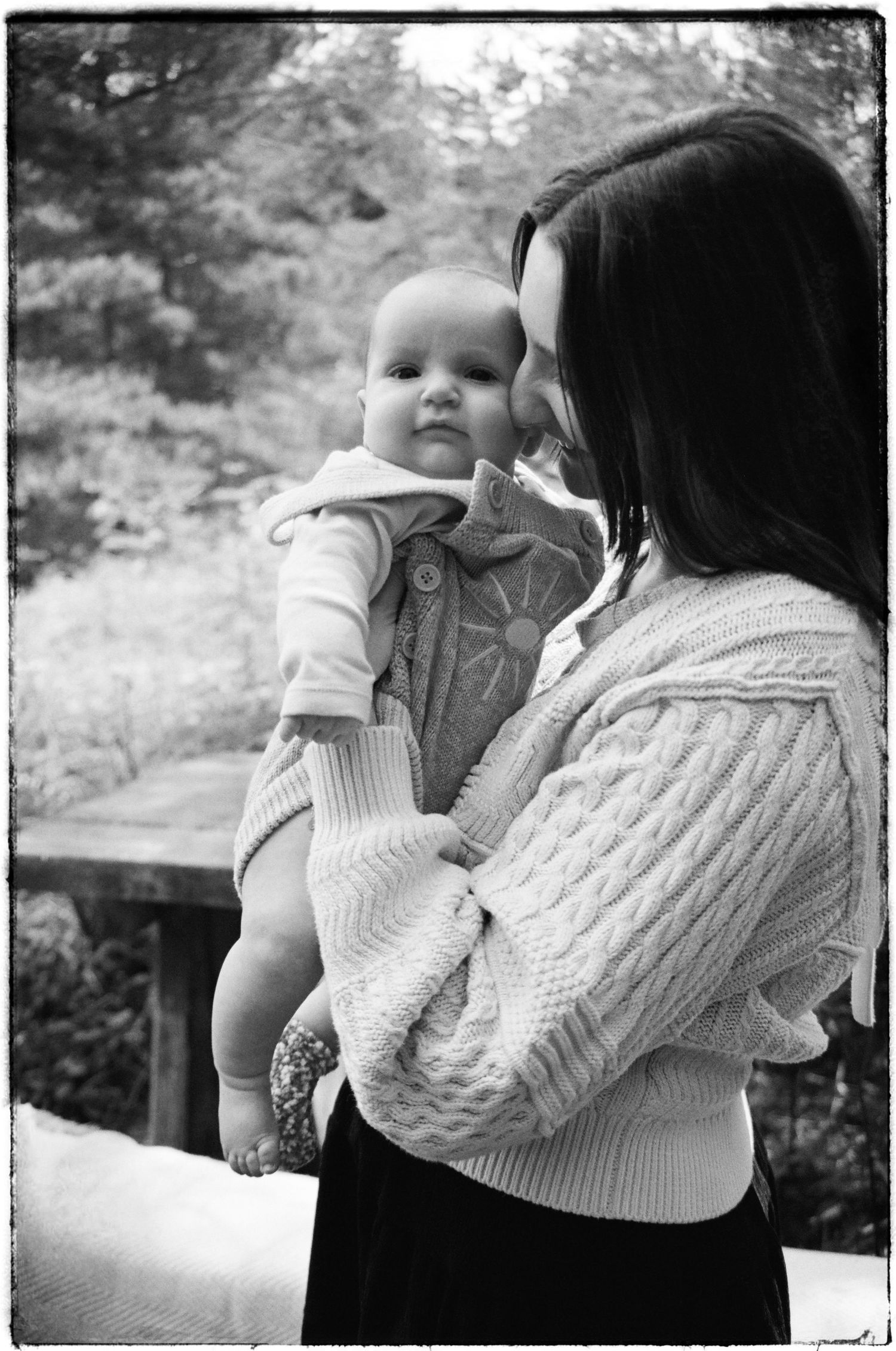 Black and white portrait showing tender moment between caregiver and baby in cable knit sweaters outdoors.