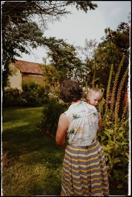 A toddler in a striped dress explores a lush garden with tall flowering plants at golden hour.