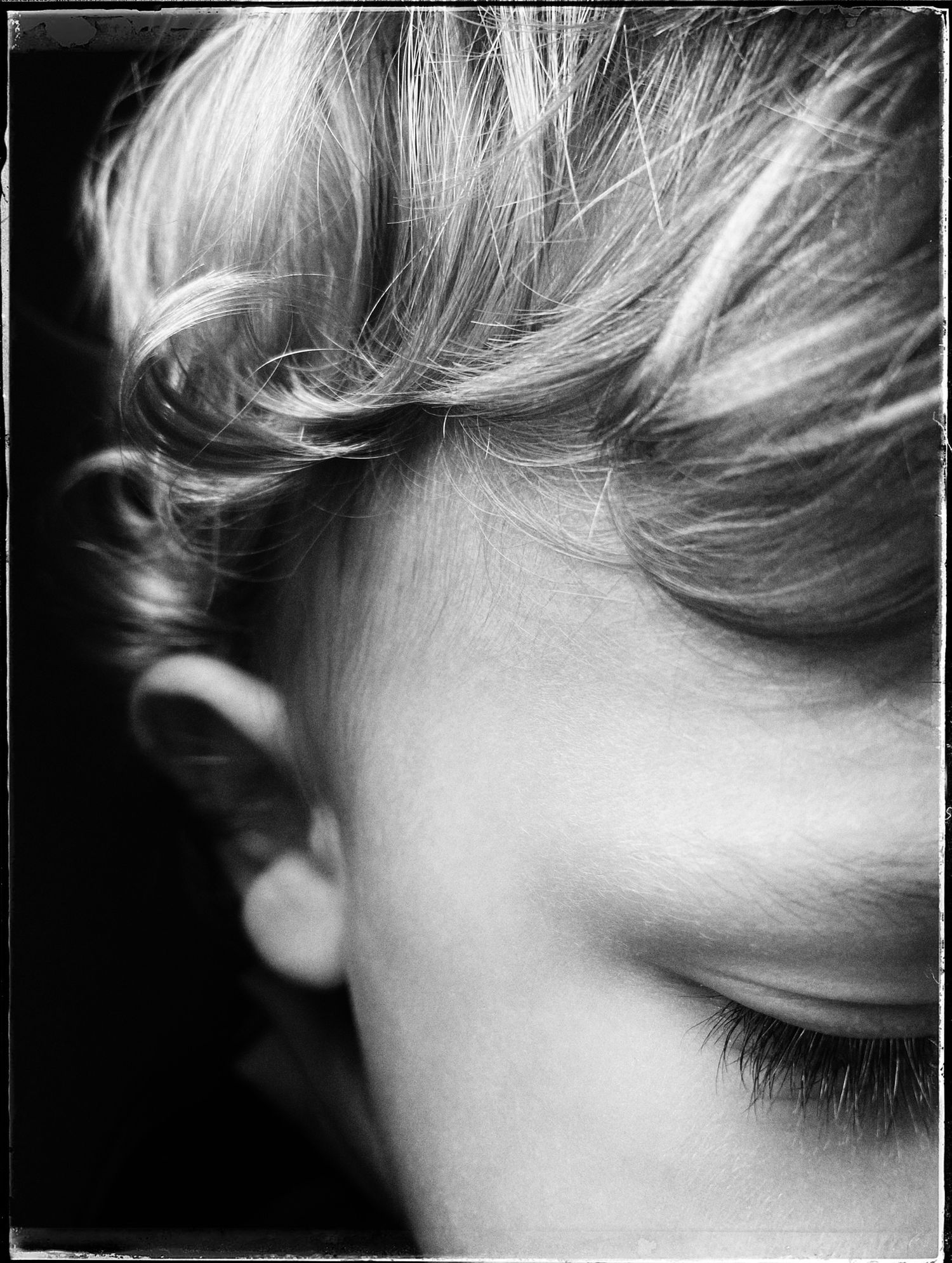 A black and white artistic close-up portrait showing wispy blonde hair and downcast eyelashes against dark background.