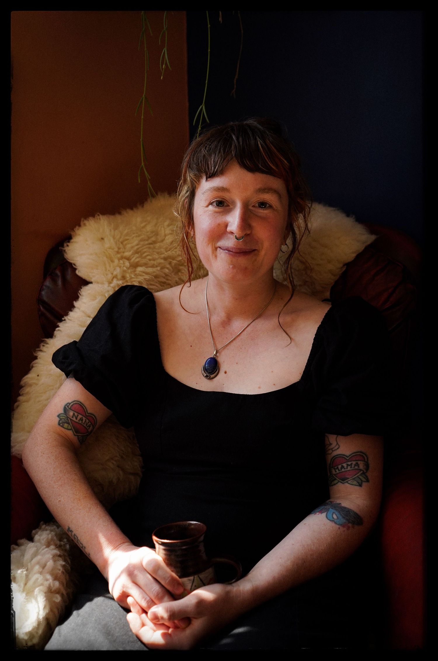 A person in black attire sits on a fur-covered chair holding a small drink, wearing visible arm tattoos and a pendant necklace.
