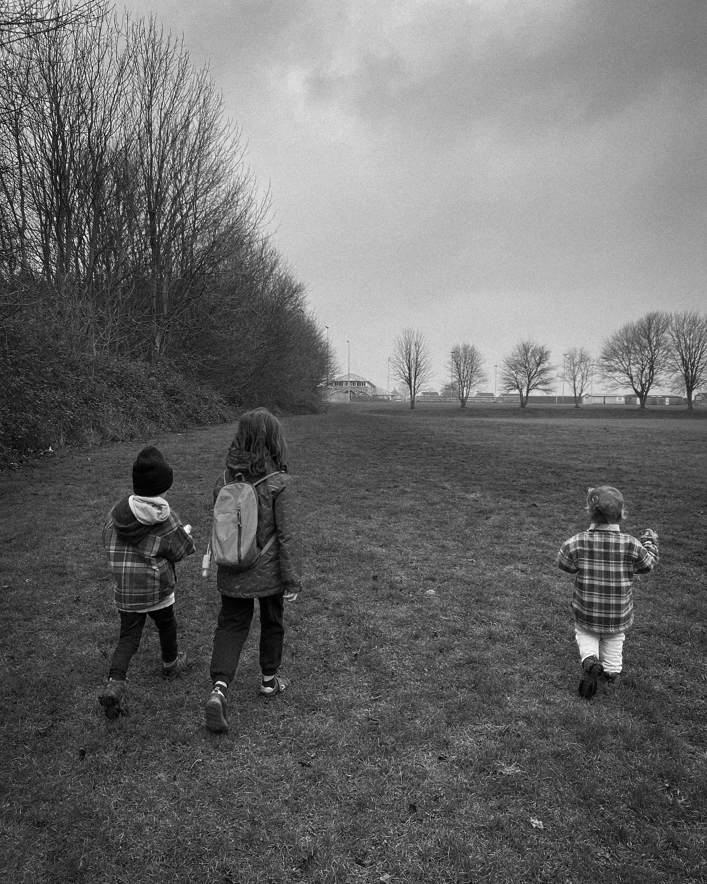 Three figures walk across a misty field with bare trees on the horizon in black and white.