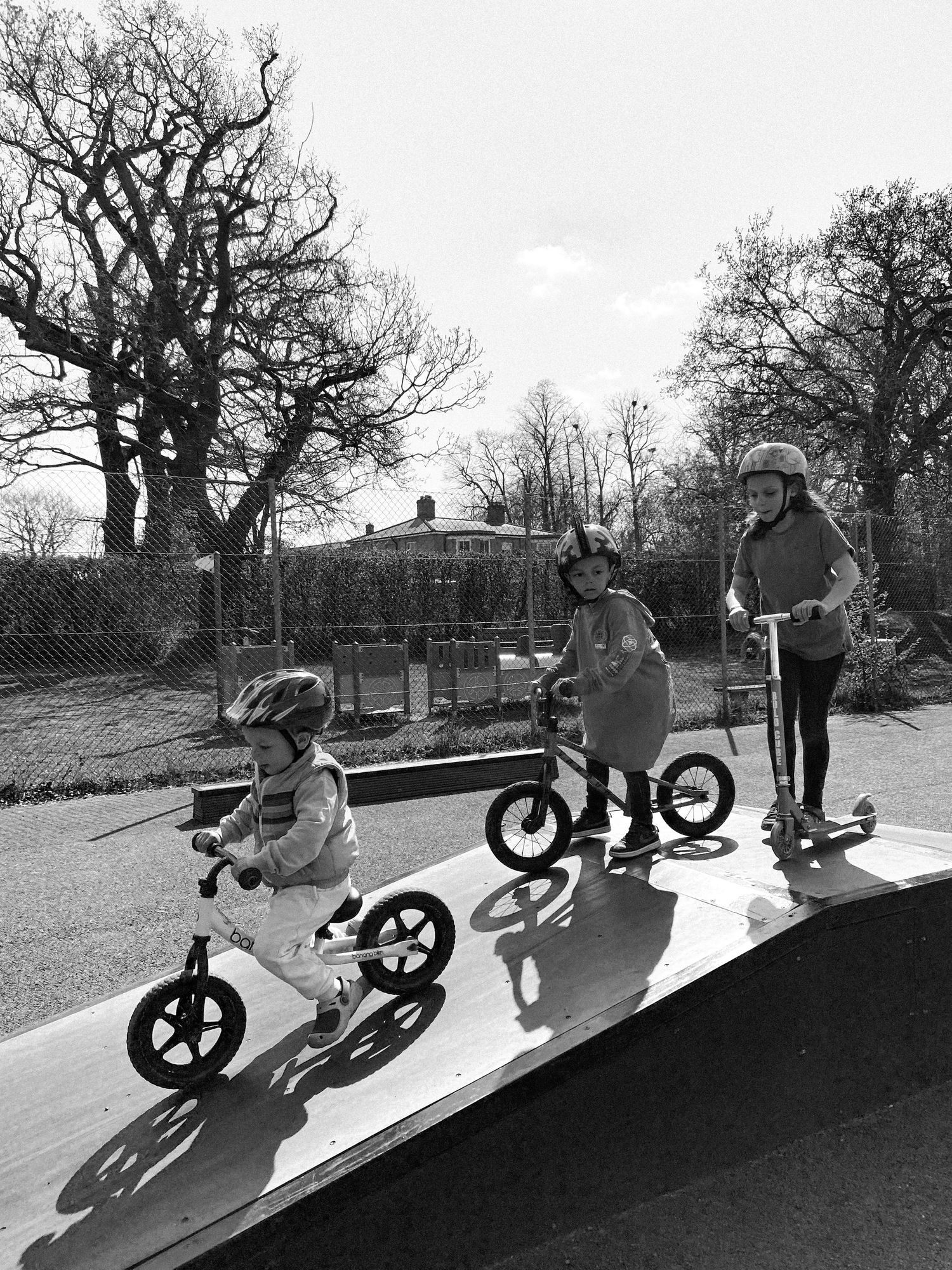 Children riding balance bikes and scooters on a ramp casting long shadows in black and white photography.