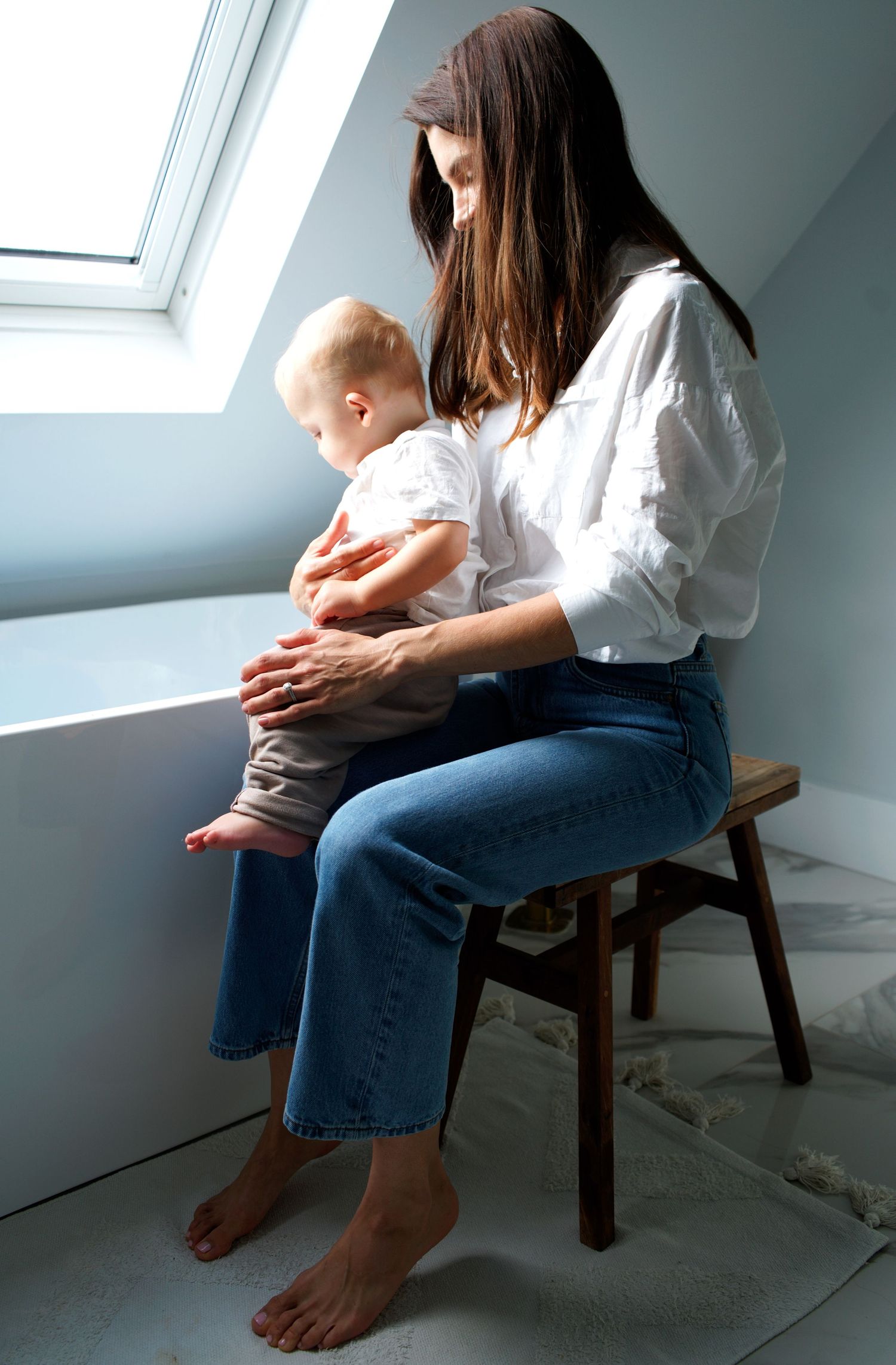 Person in white shirt and jeans sits on wooden chair in minimalist room with slanted window.