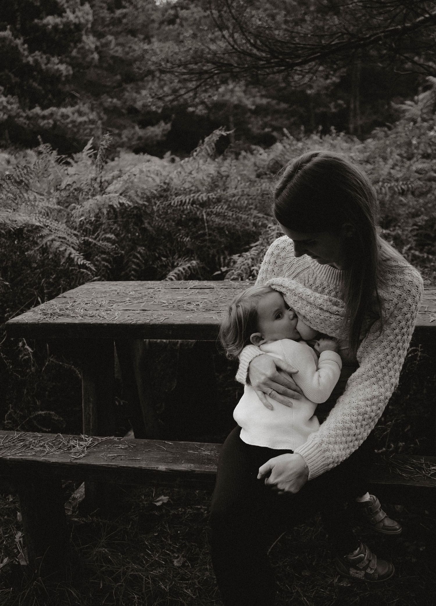 A tender black and white portrait of a parent embracing a small child outdoors among ferns and foliage.