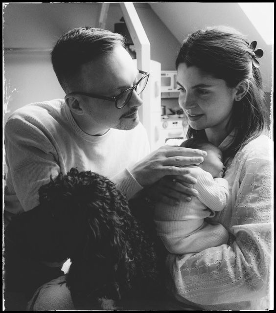 Black and white photograph of two people tenderly holding and looking at a swaddled baby in domestic setting.