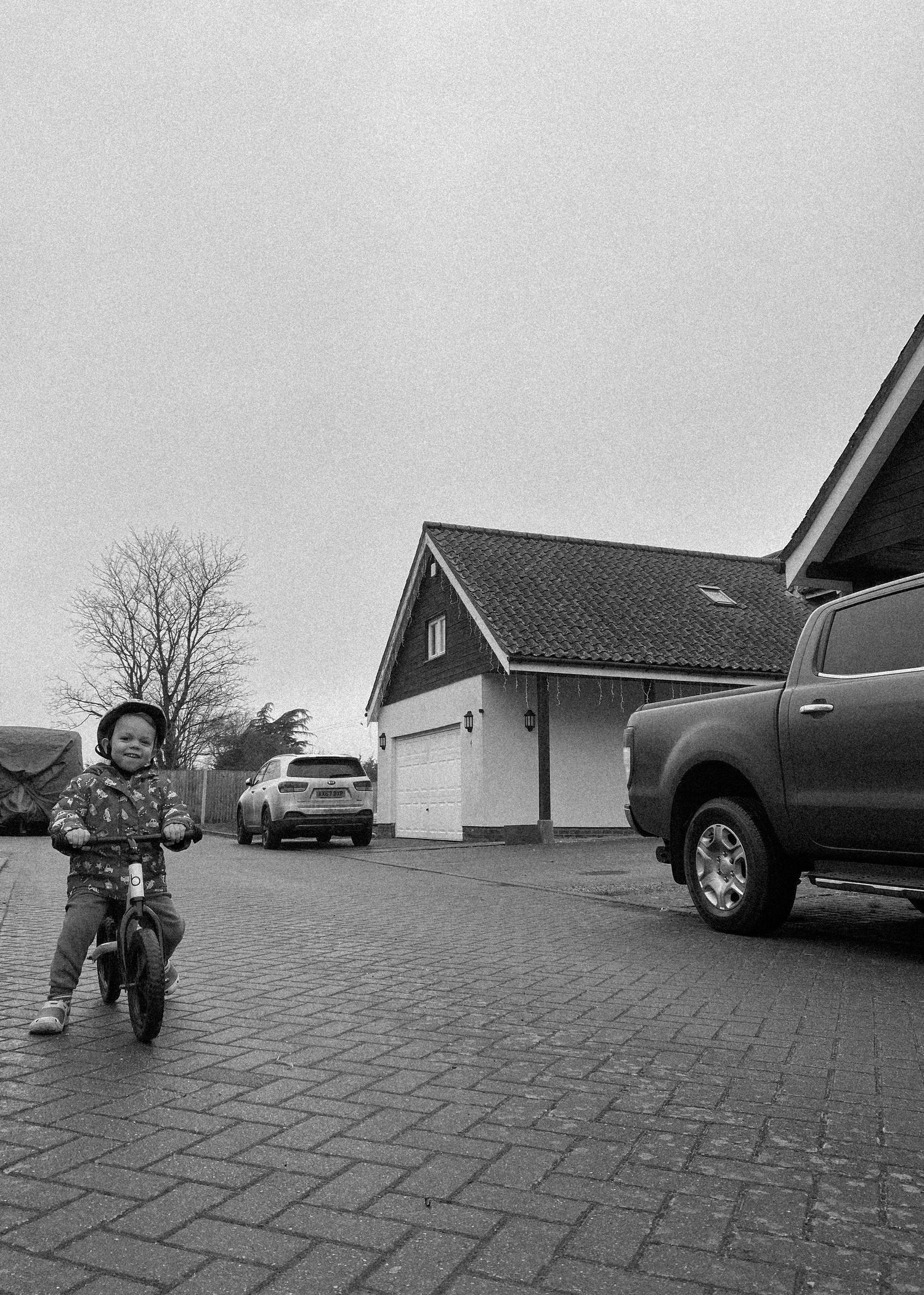 Child riding a bicycle on brick pavement with houses and parked cars in background.