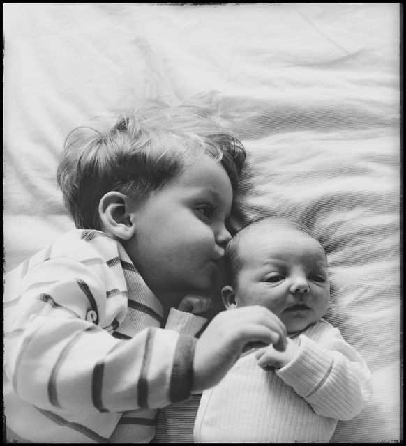 Black and white photograph shows a tender moment between two young infants lying together on soft bedding.