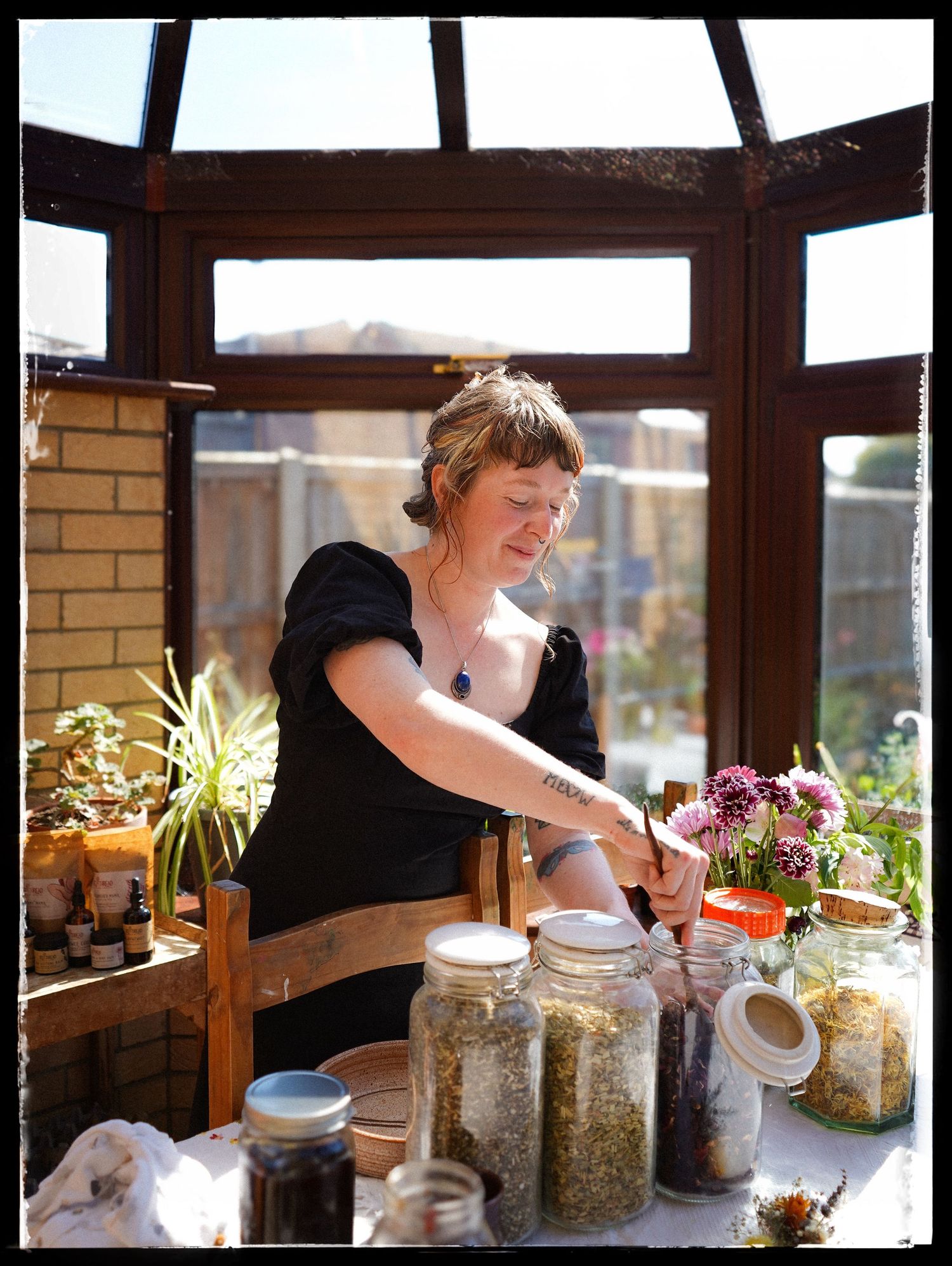 A person wearing black arranges herbs and ingredients in glass jars on a wooden counter in natural lighting.
