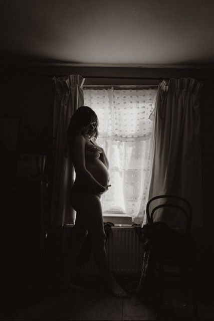 A silhouetted pregnant figure stands by a window with lace curtains in a moody black and white photograph.