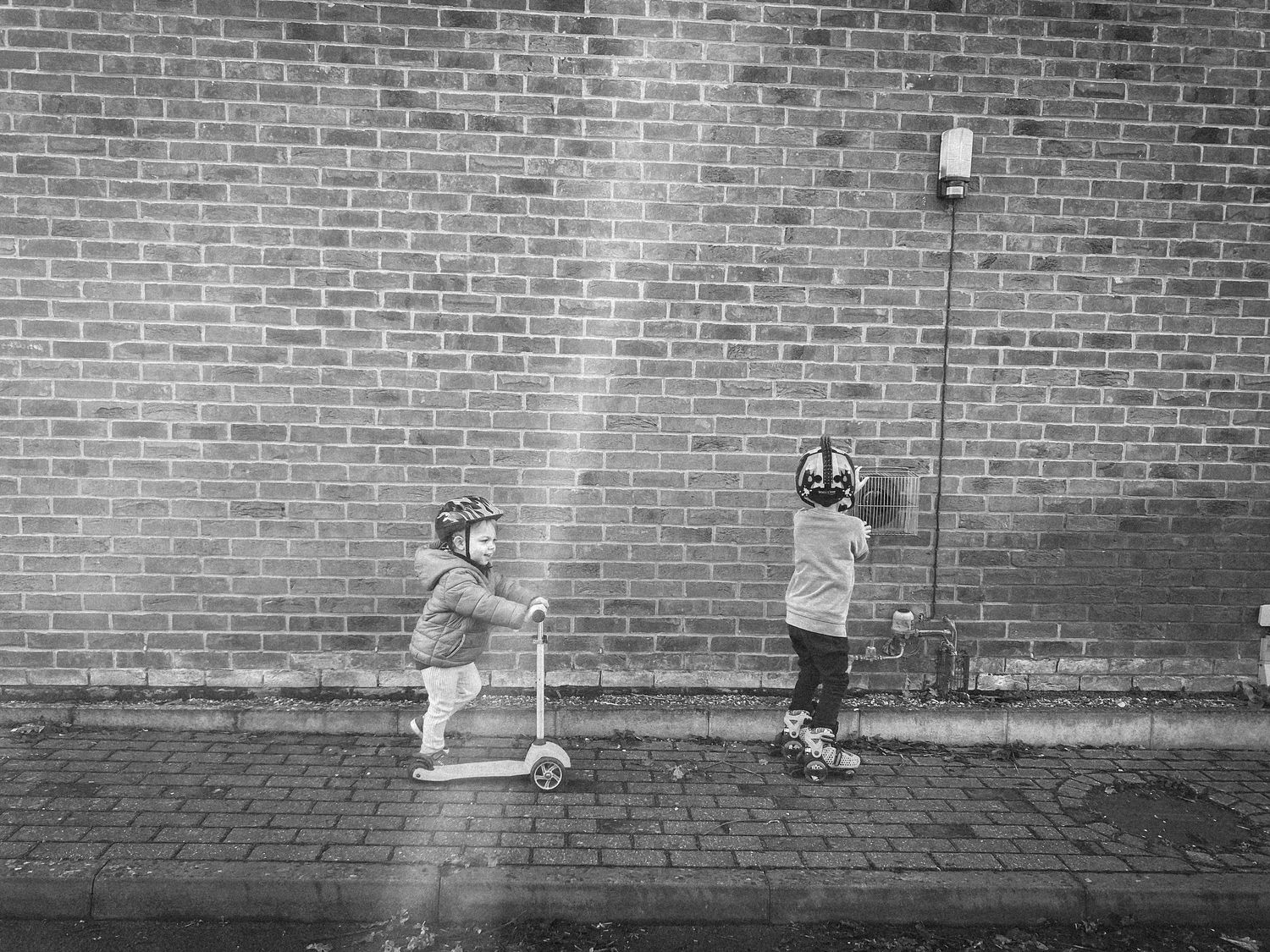 Two young riders on scooters along a brick wall in black and white.