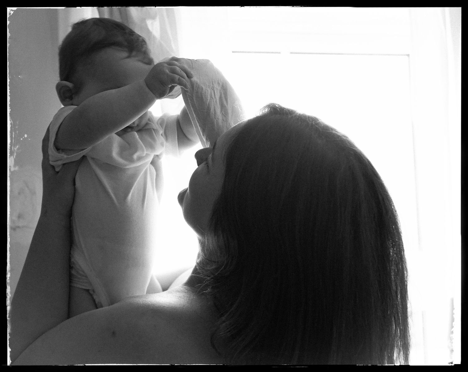A black and white intimate portrait capturing a tender moment between mother and baby in soft window light.