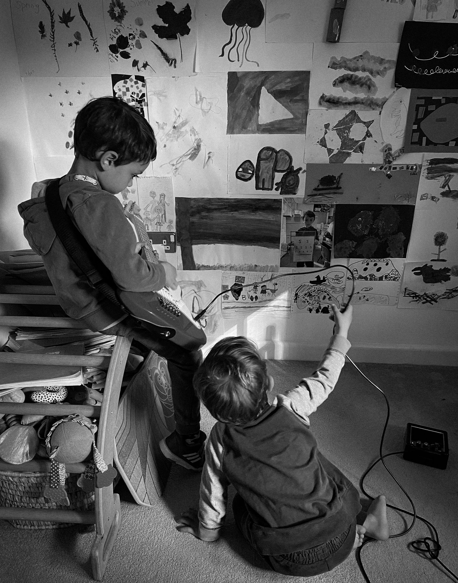 Two children sit together on the floor examining artwork on a wall covered in various drawings and sketches.