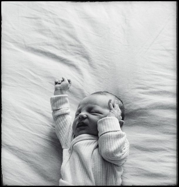 A baby wearing a white ribbed sweater lies on white bedding with their hands raised near their head.