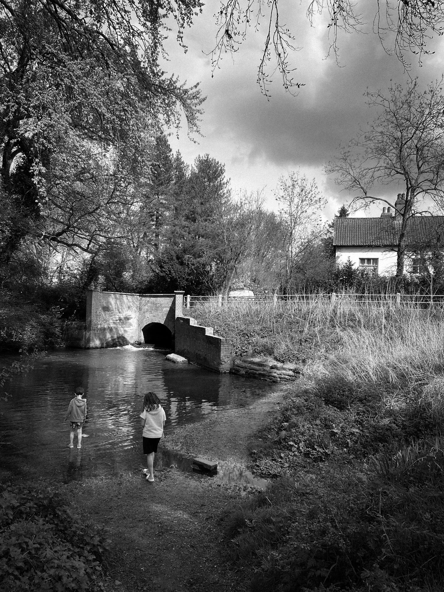 Black and white photograph of people by a small stone bridge over a stream near a rural house.
