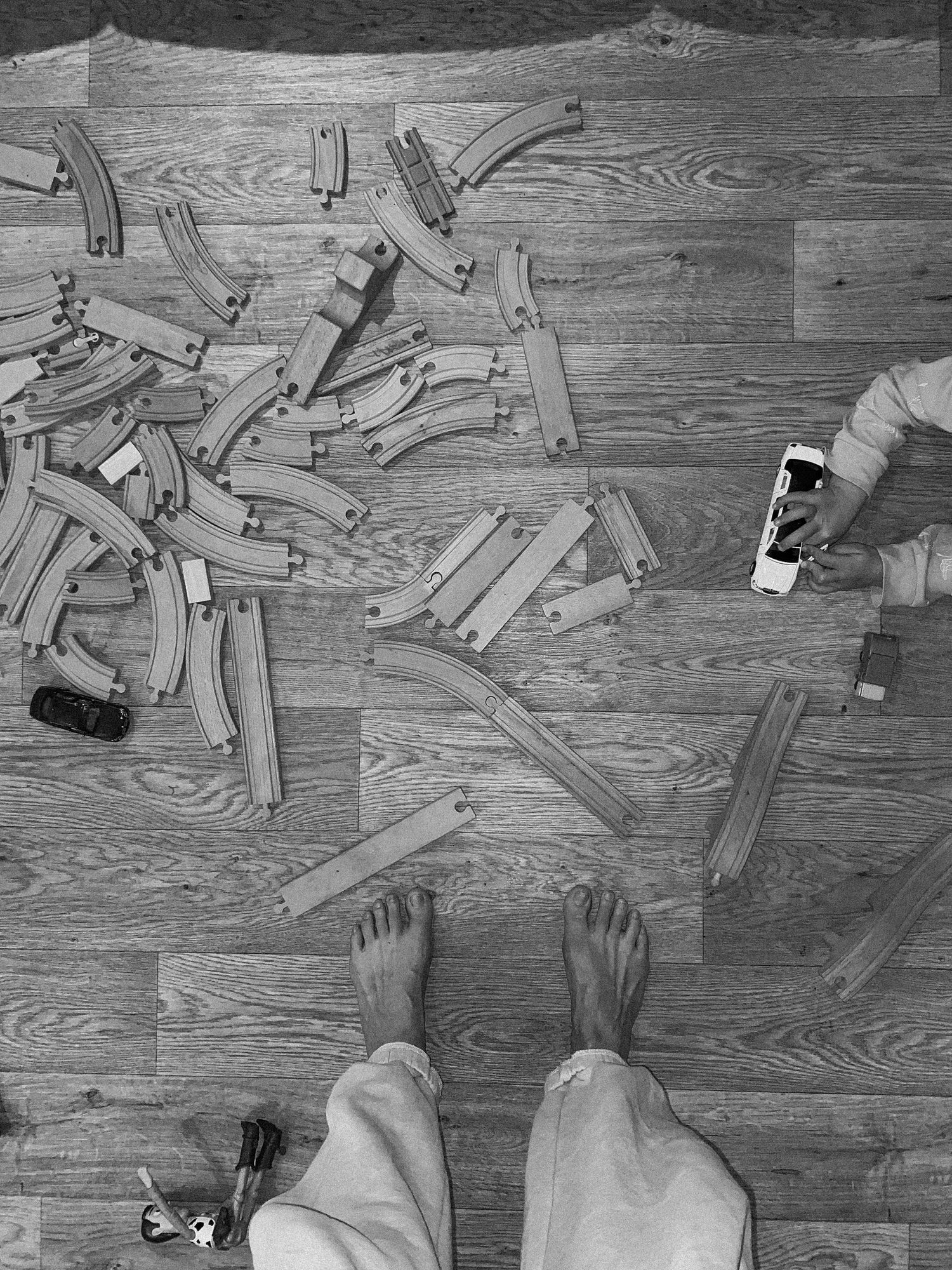 Wooden toy train tracks scattered across hardwood flooring with feet visible in the frame.
