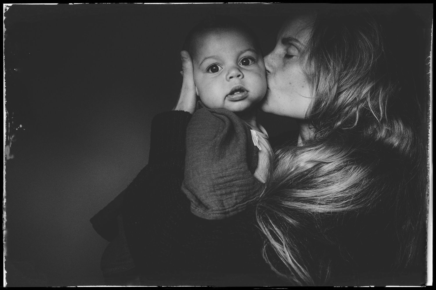 A tender black and white portrait of intimate shared moment between adult and baby against dark background.