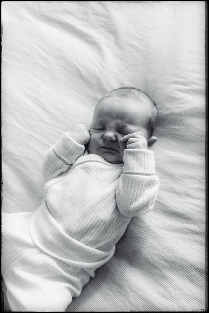 A sleeping baby in a white knit outfit rests on white bedding in black and white photography.