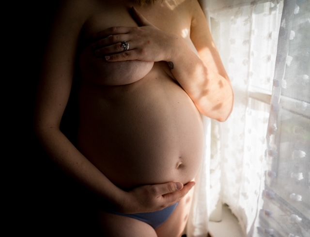 A silhouetted pregnant belly is illuminated by soft window light filtering through sheer curtains.