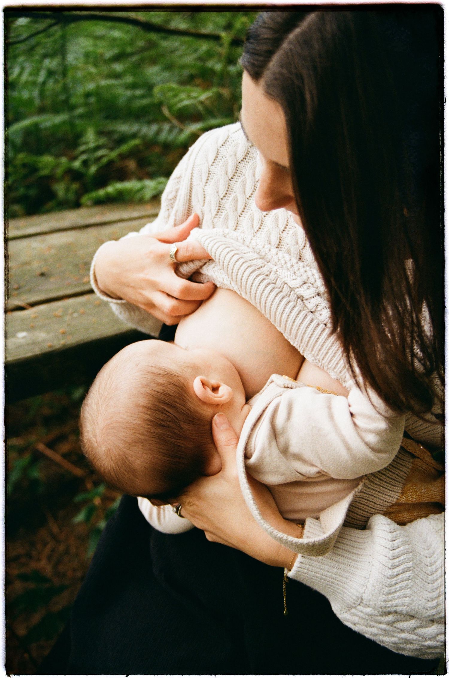 A person in a white cable knit sweater nursing a baby outdoors with green foliage in the background.