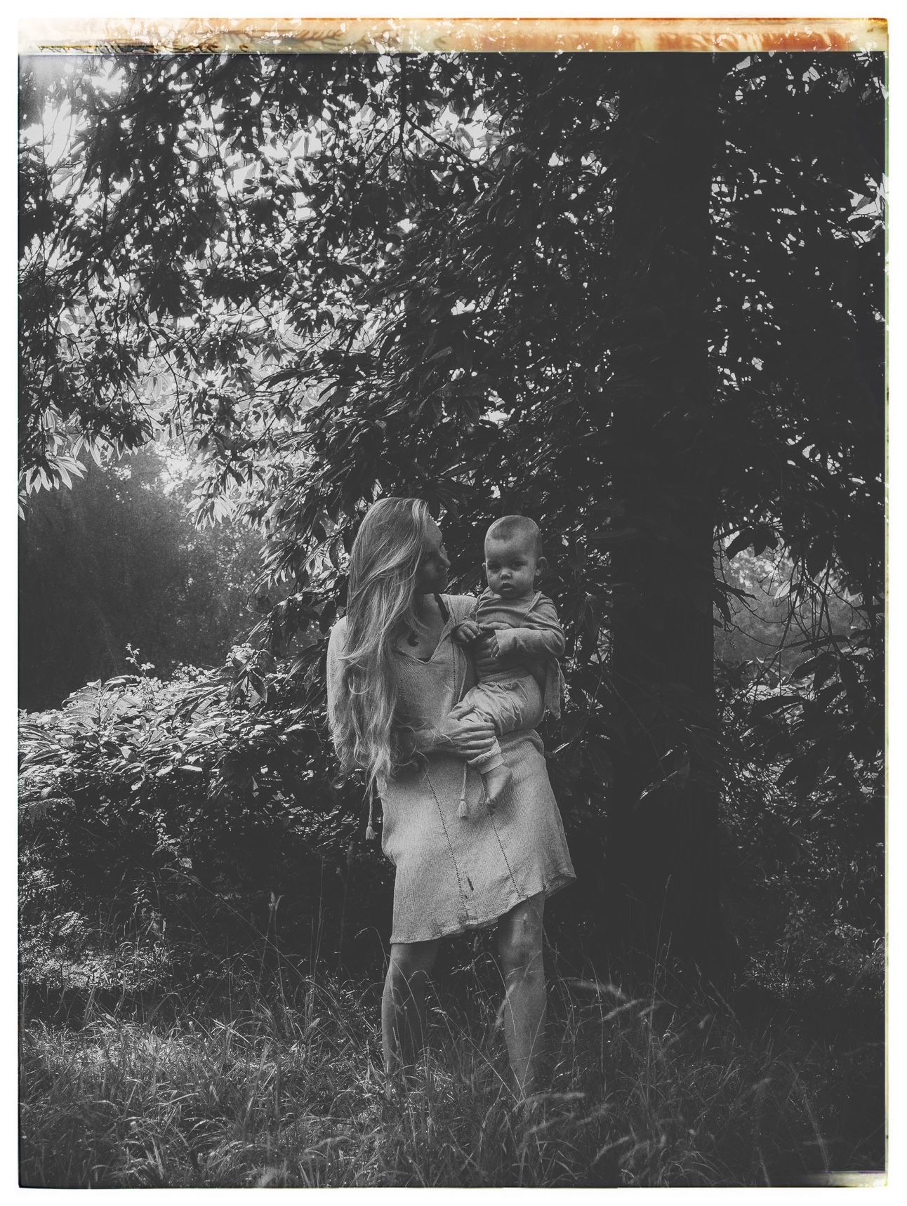Black and white outdoor portrait of parent and child standing among trees in natural setting.