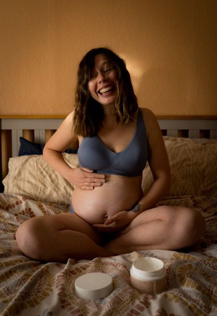 Expectant woman sits cross-legged on bed with candles in warm lighting.