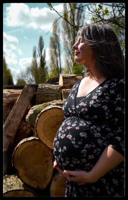 A pregnant woman in a floral dress stands near stacked logs on a sunny spring day with tall trees in the background.