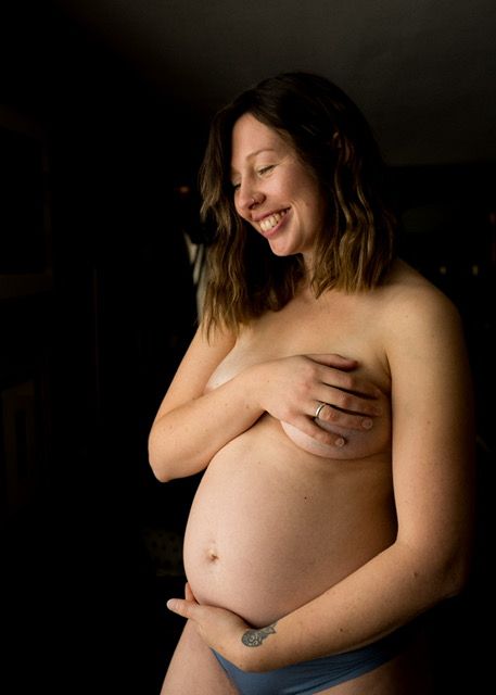 A joyful pregnant woman laughs while holding her bare belly in a dramatic dark lighting artistic maternity photo.