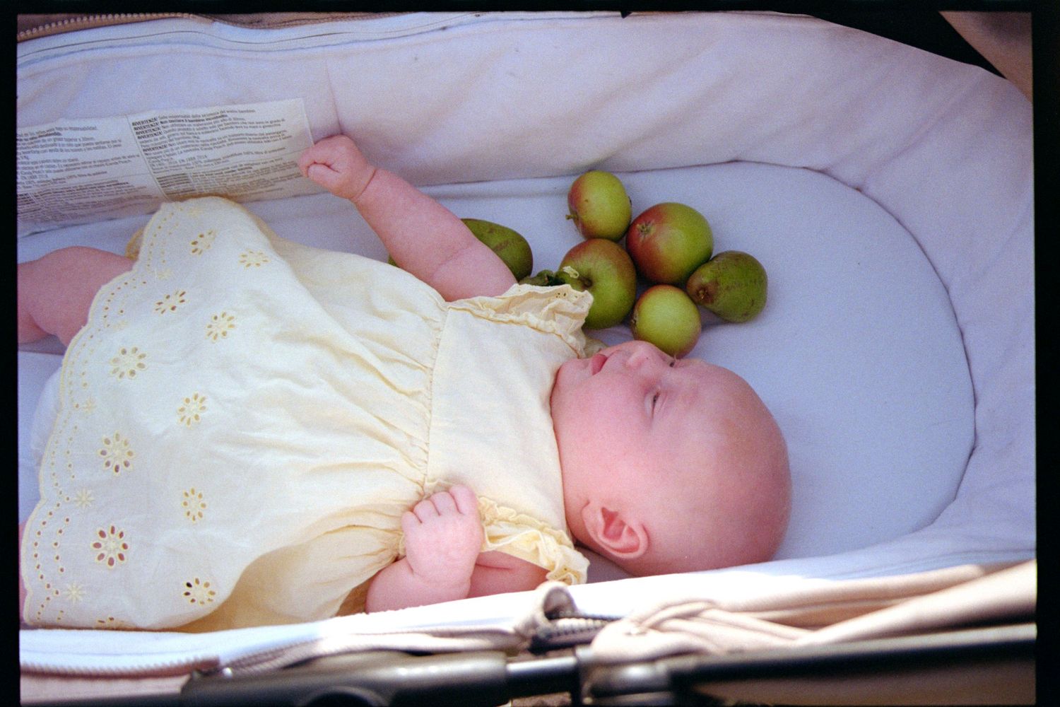 A baby lies on white bedding while someone holds fresh green olives nearby.